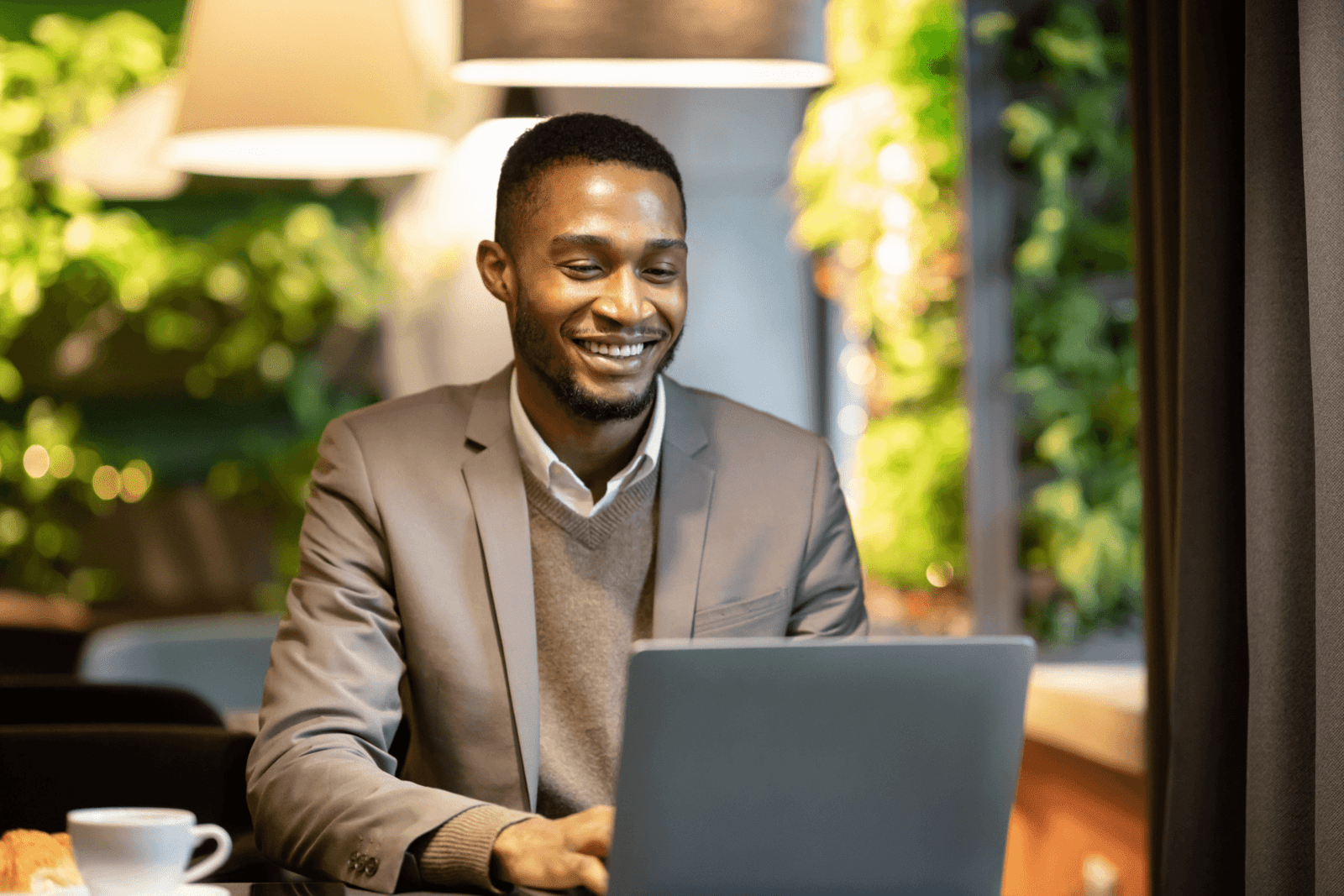 smiling professional in a suit working on a laptop in a stylish, well-lit environment with greenery in the background.