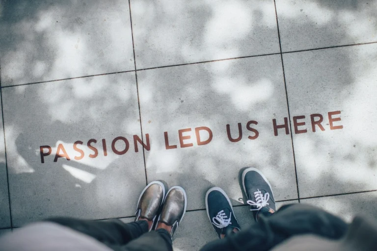 View from above of two pairs of feet standing on a sidewalk, featuring the quote "PASSION LED US HERE" printed on the ground.