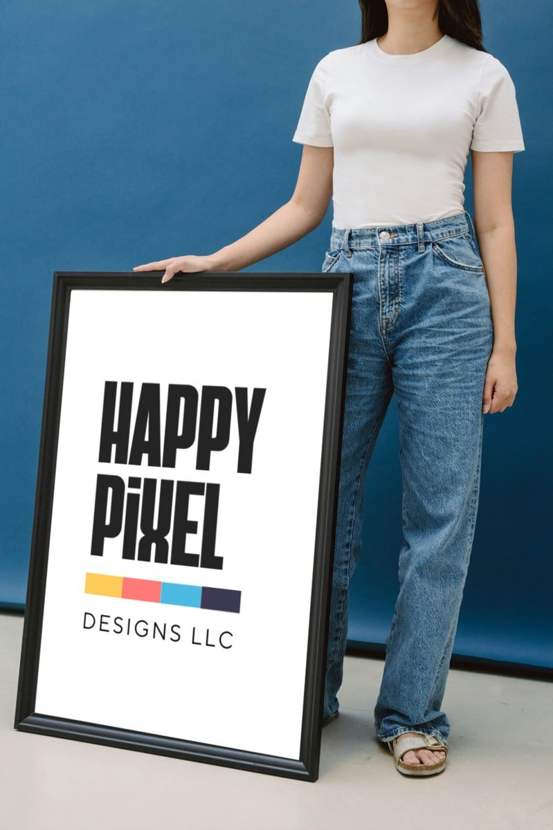 A woman in a white t-shirt holds a large black-framed poster displaying the logo for "Happy Pixel Designs LLC" against a blue backdrop.